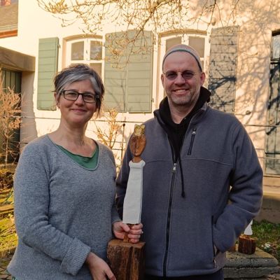 Birgit Mock und Ralf Knoblauch draußen mit grauen Pullis. Sie hält eine ca. 50 Zentimeter hohe Königinnenskulptur aus bemaltem Holz in den Händen.