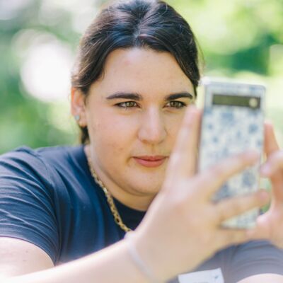 Im Grünen ist eine Frau in die Hocke gegangen und fokussiert etwas mit ihrer Handykamera.