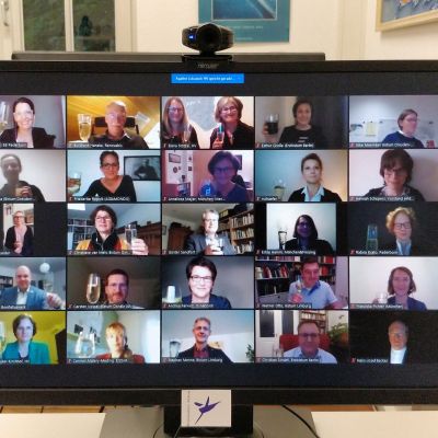 Auf einem PC-Monitor mit aufgesteckter Webcam sind 25 Gesichter von Teilnehmern der Videokonferenz zu sehen. Viele von ihnen halten ein Sektglas ins Bild ihrer jeweiligen Webcam.