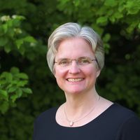 Auf dem Porträt trägt Charlotte Kreuter-Kirchhof kurze graue Haare, ein dunkles Rundhals-Oberteil und eine silberne Kette. Sie steht vor einem dichten grünem Busch und lächelt.