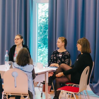Sechs eher jüngere Frauen sitzen um einen Tisch im großen Tagungssaal und unterhalten sich.