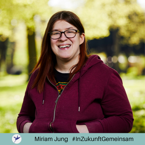 Das Bild zeigt Miriam Jung, die in die Kamera lacht. Sie trägt eine rote Sweatshirtjacke über einem schwarzen T-Shirt. Sie hat langes rotes Haar und trägt eine Brille.