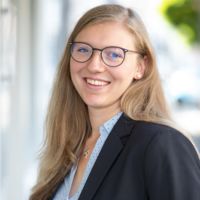 Jule Rams hat lange blonde Haare und eine Brille. Sie trägt einen schwarzen Blazer über der Bluse und lächelt.