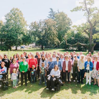 70 Frauen und drei Kinder sitzen und stehen auf einer großen Wiese im Park. Hinter ihnen sind viele unterschiedliche Bäume im Bild.
