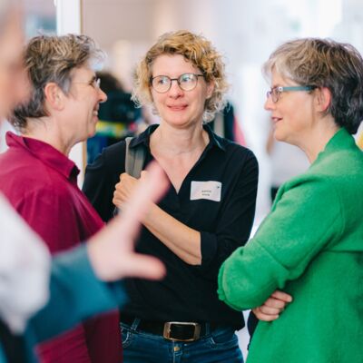 Fokussiert sind drei sich zugewandte Frauen im Gespräch. Als Rahmen des Bildes sind schemenhaft Umrisse von zwei Frauen im Vordergrund zu erkennen, sie sich zu zweit unterhalten.