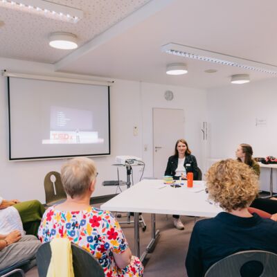 Acht Frauen sitzen um eienn Tisch und sehen sich einen TEDx-Talk an.