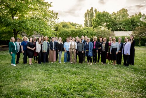 26 Frauen und ein Mann stehen im Grünen vor einer dramatischen Sonne-und-Wolken-Kulisse.