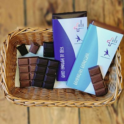 Zwei Tafeln von dunkler und Milchschokolade mit den Logos vom Hildegardis-Verein und dem Synodalen Weg in einem Körbchen. Auf einer Tafel steht "gemeinsam genießen" und "auf der anderen "good enough to try".