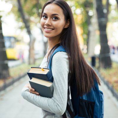Lächelnde, dunkelhäutige Studentin mit langen braunen Haaren. Sie trägt einen Rucksack auf dem Rücken und Bücher im Arm.